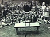 Groupe d'enfants.  Une enfant contorsionniste sur une table fait un spectacle à l'extérieur devant parents et enfants qui applaudissent ; acrobatie