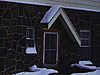 Une dame sort du chalet, un homme pellette l'entrée très enneigée da la maison (il neige); neige abondante