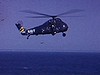 Hélicoptère (NAVY) en vol qui atterrit sur le porte-avion; des gens circulent sur le pont; l'hélicoptère décolle (aileron d'avion avec tête de mort)