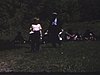 Famille nombreuse de tous les âges, assis dans l'herbe en pique-nique. Certaines femmes âgées assises dans des chaises. Partie de fers sur le terrain, hommes en chemises, bretelles cravates et vestons