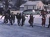 Gens de tous âges patinent sur une étendue glacée au coeur d'un village enneigé, course de patins de jeunes femmes et adolescentes, suivie de jeunes hommes et adolescents et garçonnets en patins