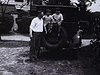 Enfants assis sur le pneu de secours à l'arrière de l'automobile  très ancienne; enfants embrassent le père; puis ils sont dans l'auto, au volant; le père frotte le pare-chocs (beaux plans)