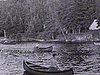 Des enfants en canots sur un lac, près d'un chalet en bois rond; chien sur le quai; canots en action