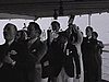 Défilé sur le pont du bateau des passagers qui tiennent des bouteilles d'alcool vides à bout de bras (chantent et marchent): groom en avant, ils portent un signet à leur veste.