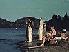 Bord de l'eau; baignade d'enfants, adolescents et adultes près du quai; chaloupe et prêtre à bord de la chaloupe; il en descend