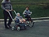Peite fille arrive vers la caméra à grande vitesse, sur son tricycle, sur le trottoir (beau plan) - puis père avec le bébé en carosse et la petite en tricycle