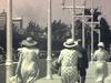 Dames avec chapeau vues de dos marchent sur le large trottoir d'une rue passantes aux États-Unis (années 30)