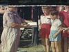 Grand-mère tend une boite de friandises aux enfants. Les femmes posent au grand vent, avec les enfants devant le lac. Sur le bord du lac, sur la pointe, un drapeau Union Jack flotte au vent