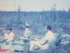 Une réunion de famille dans un terrain vague en Abitibi, assez désertique, des chaises longues dans une forêt de conifères, de nombreuses personnes, des enfants bien habillés, vestes de laine, des gens assis par terre ou sur des bûches, le temps semble as