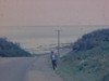 Femme marche sur le bord de la route en Gaspésie, paysage, bord de mer, île Bonaventure, rocher Percé, bateaux de pêche amarrés au quai sur mer agitée - 1966