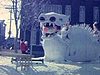 Personne devant monument historique et à côté de deux lutins et un renne descendent une colline, sculpture géante du Bonhomme Carnaval, une femme pose devant un moulin '' L'ami du Québec'', pyramide de glace, parade, fanfare, plan de sculptures sur glace