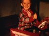Des enfants avec leurs cadeaux de Noël: un garçon dans une voiture à pédales, une fillette avec une poupée dans une chaise haute, elle montre un jouet à sa mère (sombre)