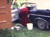 Maison de campagne, une femme nettoie une automobile, tracteur près du garage, plusieurs carcasses d’autos sur le terrain, femme et enfants devant. un camion de constrution passe sur la route