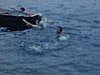 Baignade dans un lac, trois personnes se tiennent accrochées à une chaloupe, un homme donne un coup de pied dans les airs hors de l’eau - 1955