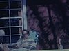 Famille au chalet, des femmes assises sur la rampe de la galerie, un homme étendu dans une chaise pliante lit un livre, journée ensoleillée - fin des années 60