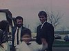 Deux hommes en habit et cravate regardent la caméra, un garçon en habit de communiant se met devant eux et pose, fait des singeries, son petit frère marche vers la caméra en se grattant la tête