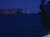 A bord d’un bateau en mouvement, profil de femme aux lunettes fumées et foulard sur les cheveux qui regarde le rocher Percé, Gros plan sur la rive de Percé au premier plan, toit de la cabine du bateau, Homme avec casquette de capitaine qui discute.