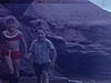 Deux enfants au bord de l’eau sur les rochers, terrain de camping, une famille essaie de faire à manger près de la tente, garçon allume poêle Coleman, un grosse flamme en sort. une fille mange un blé d’inde