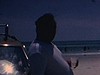 Des gens en maillot de bain réunis près des autos, un homme bedonnant court et traverse un ruisseau, un couple s’embrasse, enfants assis sur des trippes saluent la caméra et posent, deux hommes discutent et prennent une bière sur le capot d’une auto, un a