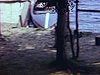 Famille au bord de l’eau, sur la plage, un homme sur une chaise adirondack parle avec une fillette qui joue à la poupée, chaloupe sur la plage 