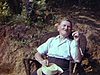 Un homme dans une chaise en rotin se prélasse au soleil, famille dans une chaloupe, une femme pagaie pour ses trois enfants et un homme âgé - chalet