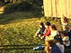 Fête de famille à la campagne, des enfants assis sur l’herbe près de la grange, des adultes autour les regardent, coucher de soleil, un garçon joue avec une tondeuse en plastique Fisher Price, des hommes dans la boite d’un camion qui roule dans le champ