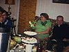 Table remplie de nourriture pour le réveillon de Noël. Deux femmes discutent, un petit garçon s’approche de la table, deux hommes au bout et l’un d’eux prend une bouchée de sandwich et donne l’autre moitié à son ami, ils rient beaucoup. des gens assis sur