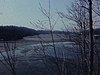 Printemps, lac en face d’un chalet qui dégèle, un homme et une femme qui filme marchent vers la caméra, un peu de neige fond