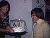 Anniversaire d’un homme - il souffle la bougie géante sur son gâteau, la femme dépose le gâteau et lui donne un cadeau, la famille autour le regarde, un couple danse - sous-sol, look années 70, moustache - tout le monde s’embrasse