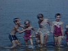 Deux petites filles en costume de bain tiennent un bébé garçon par la main, ils sont dans l'eau et marchent vers la caméra, il sourit à la caméra, une petite fille marche, les enfants s'amuse dans l'eau et marche en ligne vers la caméra, année une petite 