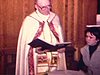 Baptême d'un bébé, le curé met le liquide sur le front de l'enfant, il récite la prière et tout le monde est autour du bébé, les témoins signent le livre, années 80