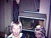 Adultes dans le salon boivent de l’alcool, les enfants dansent et s’amuse (Noël 1970) dans la cuisine: des gens dansent, une femme danse en coupant un pâté à la viande (rigolo)