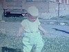 Bébé garçon en salopette et une petite casquette marche dans l’herbe, des enfants dans la rue font de la bicyclette, une femme enceinte dehors avec eux, la grande soeur tient son petit frère dans ses bras - 1964