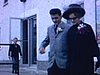 Un homme et une femme en beaux habits sortent d’une salle de réception et se dirigent vers une automobile des années 50, la famille les suit. Une femme prend des phototo lorsqu’ils entrent dans l’auto