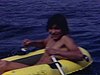 Un adolescent sur un bateau gonflable rame sur la rivière, une fille sur un matelas gonflable salue la caméra, des gens dans un bateau ramassent les skis nautiques qui flottent