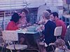 Famille à table à pique-nique, près de la roulotte en camping, ils trinquent, verres de vin, les enfants mangent sur une petite table près d’eux